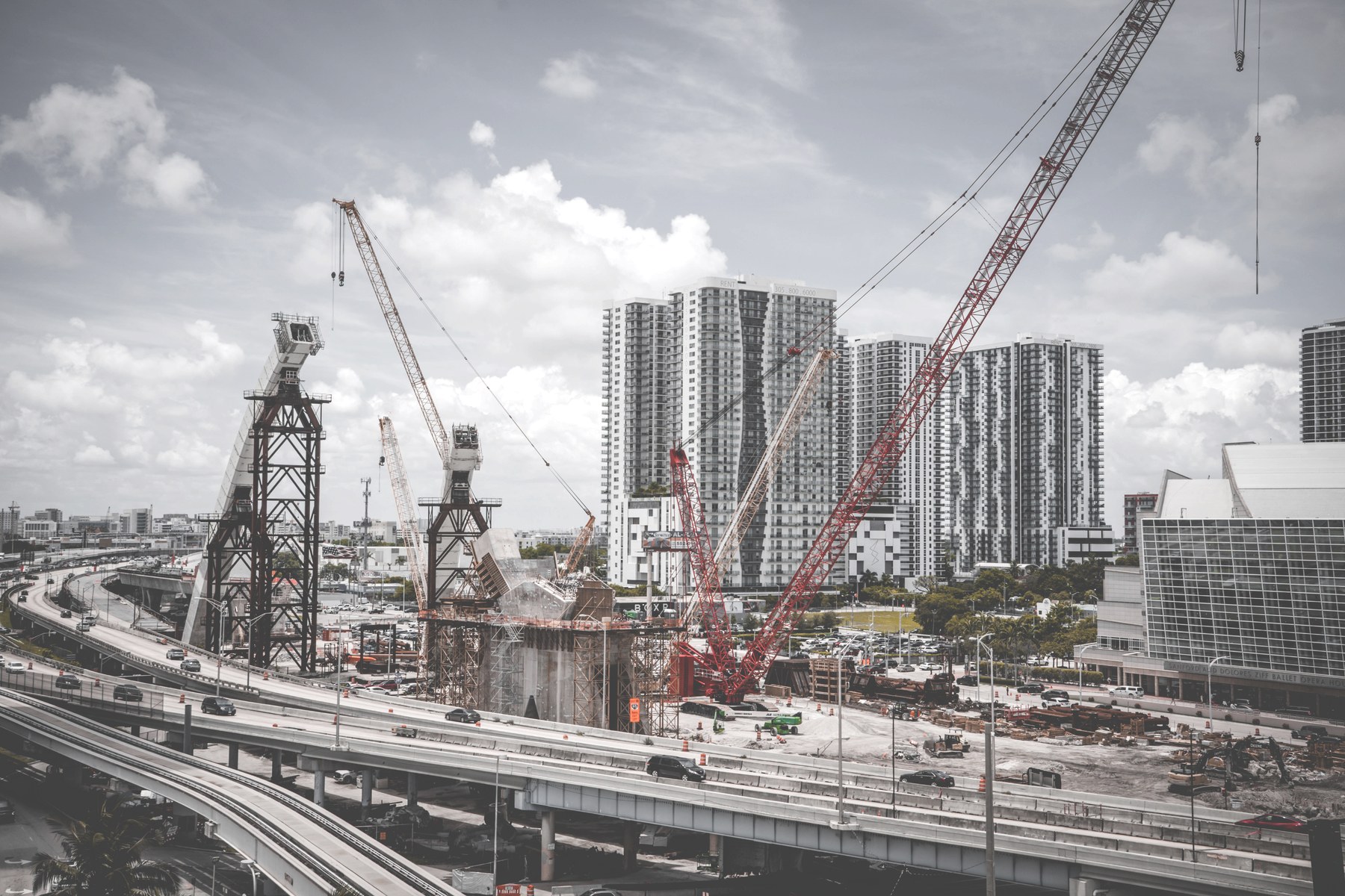 A downtown construction site with a red crawler crane raising a column, an active elevated highway, and high-rise towers under a bright overcast sky